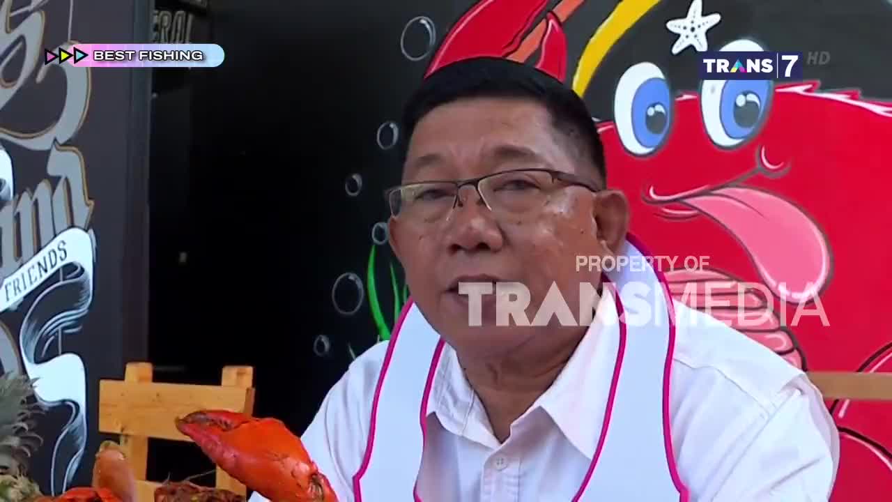 A man in glasses is speaking, with a bright red cartoon crab behind him. The Trans7 logo is visible, indicating this is likely a segment from Indonesia.
