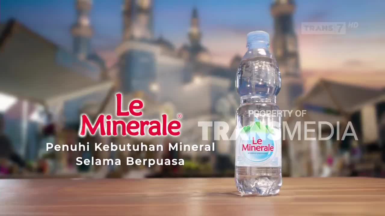A bottle of Le Minerale stands on a wooden surface. Behind it, blurred buildings with minarets suggest a mosque, and the Trans7 logo is visible.