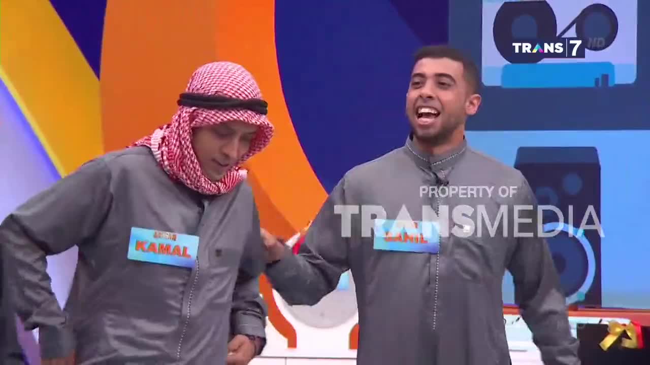 Two men in grey robes stand on a colorful stage, one with a red and white keffiyeh on his head. The man on the right grins widely, his mouth open as if speaking or laughing. A Trans 7 logo is visible in the background, suggesting this is a broadcast from Indonesia.
