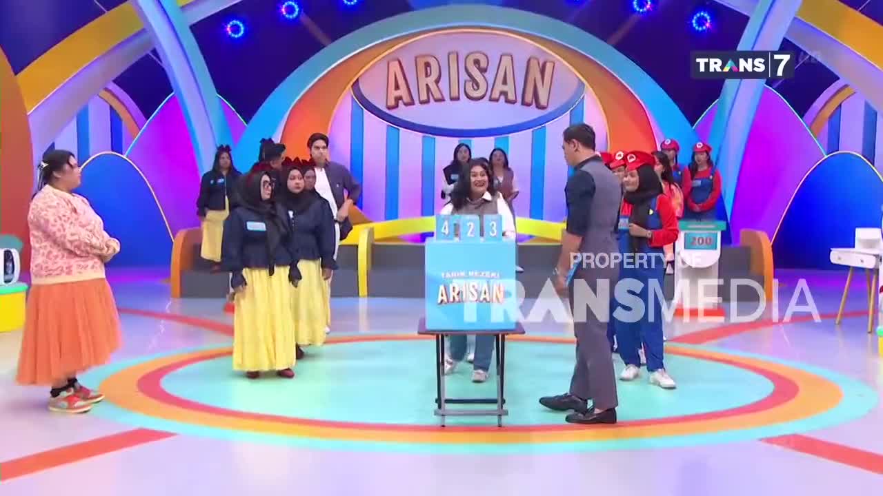 A woman in a bright orange skirt stands on the left, facing a group of women in matching blue jackets and yellow skirts. A man in a dark vest and grey pants stands center, looking towards a blue box with numbers on it. The "Arisan" logo hangs above the stage.