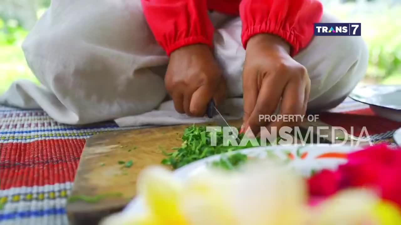 Fingers expertly mince fresh herbs on a wooden board. The vibrant red of a Trans7 shirt stands out against the pale fabric of a sarong.