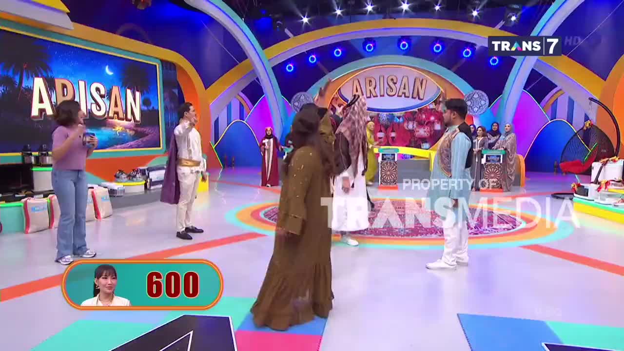 A woman in a flowing brown robe stands center stage on the Trans7 set, facing away from the audience. To her left, a man in a white prince-like costume gestures upwards, while a woman in a purple top and jeans holds a drink.