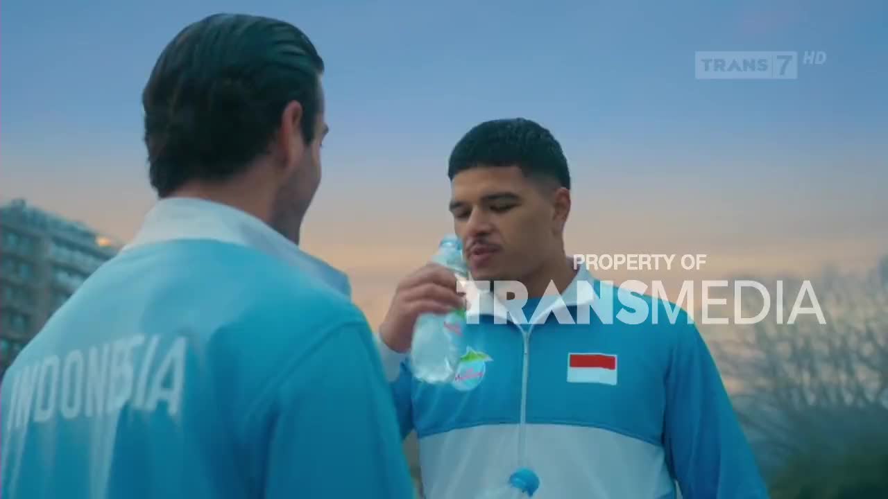 A young man in a blue tracksuit with the Indonesian flag on his chest takes a drink from a water bottle. His teammate, also in team gear, stands nearby, his back to the camera. The Trans7 logo is visible in the upper right corner.