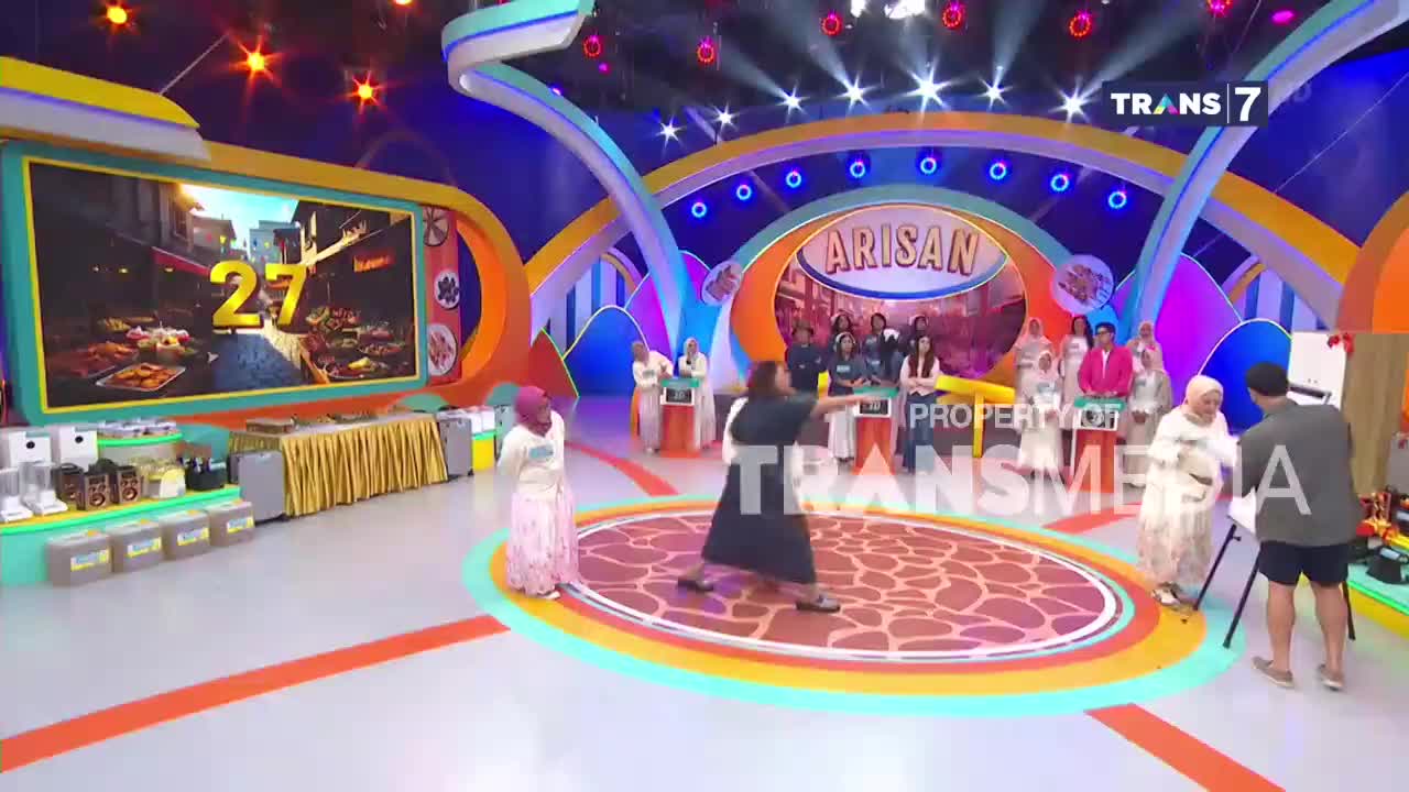 A woman in a black dress spins towards a woman in a white hijab on a game show stage. The large screen behind them displays a street scene with the number 27. A woman in a black dress spins towards a woman in a white hijab on a game show stage. The large screen behind them displays a street scene with the number 27.