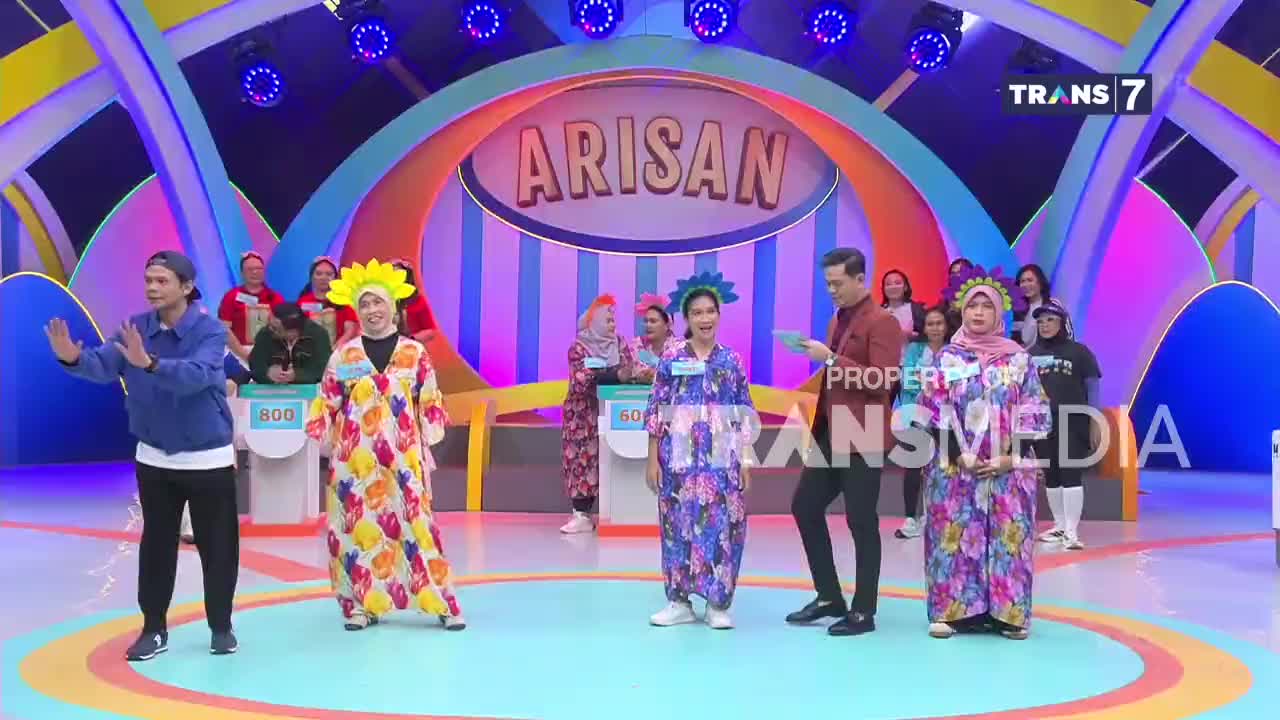 The host, dressed in a maroon jacket, stands on stage with a contestant in a floral dress. Several other participants in colorful attire wait behind them as the "Arisan" game show unfolds. The host, dressed in a maroon jacket, stands on stage with a contestant in a floral dress. Several other participants in colorful attire wait behind them as the "Arisan" game show unfolds.