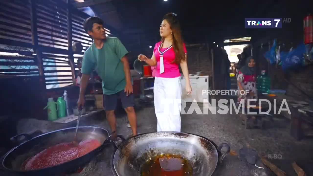 A man stirs a large pot of bright red liquid over a fire while a woman in a pink shirt and white pants watches. Behind them, another person tends to a smaller pan of oil. A man stirs a large pot of bright red liquid over a fire while a woman in a pink shirt and white pants watches. Behind them, another person tends to a smaller pan of oil.