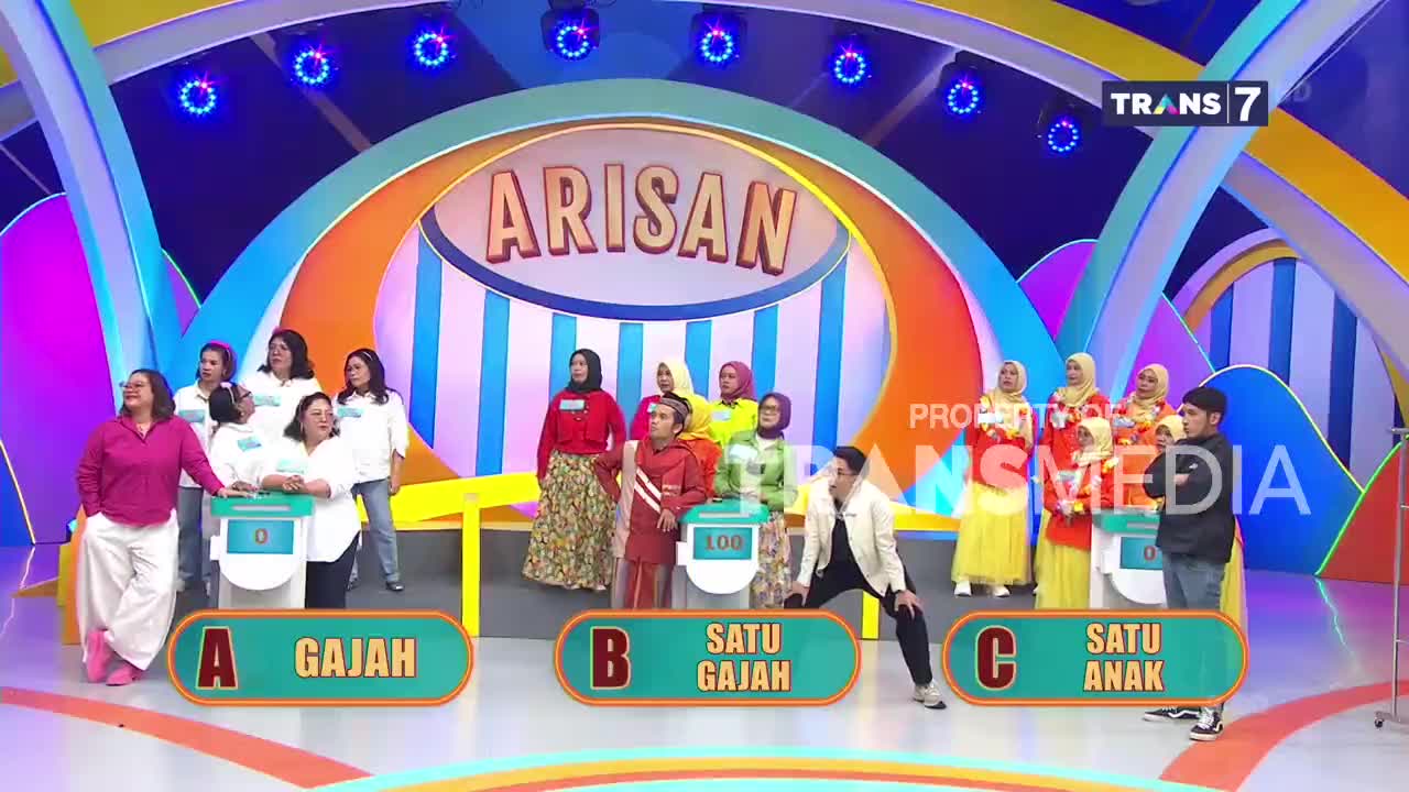 A group of women stands on a brightly lit stage, some in white shirts, others in colorful outfits, facing a game show setup. A man in a white jacket crouches low in the foreground, looking towards the contestants. A group of women stands on a brightly lit stage, some in white shirts, others in colorful outfits, facing a game show setup. A man in a white jacket crouches low in the foreground, looking towards the contestants.