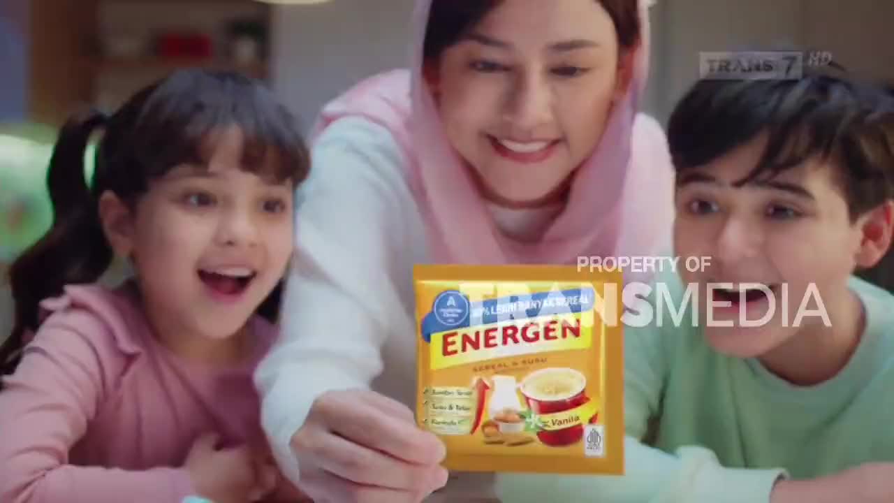 A woman holds up a packet of Energen cereal for two excited children. The Trans7 logo is visible in the background. A woman holds up a packet of Energen cereal for two excited children. The Trans7 logo is visible in the background.