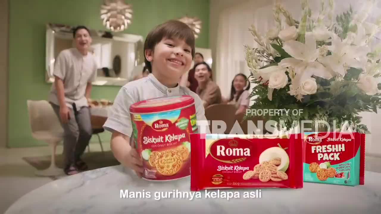 A boy grins, holding up a large red tin of Roma Biscuits Kelapa. Behind him, a family laughs and a man in a light-colored shirt bounces on the balls of his feet. A boy grins, holding up a large red tin of Roma Biscuits Kelapa. Behind him, a family laughs and a man in a light-colored shirt bounces on the balls of his feet.