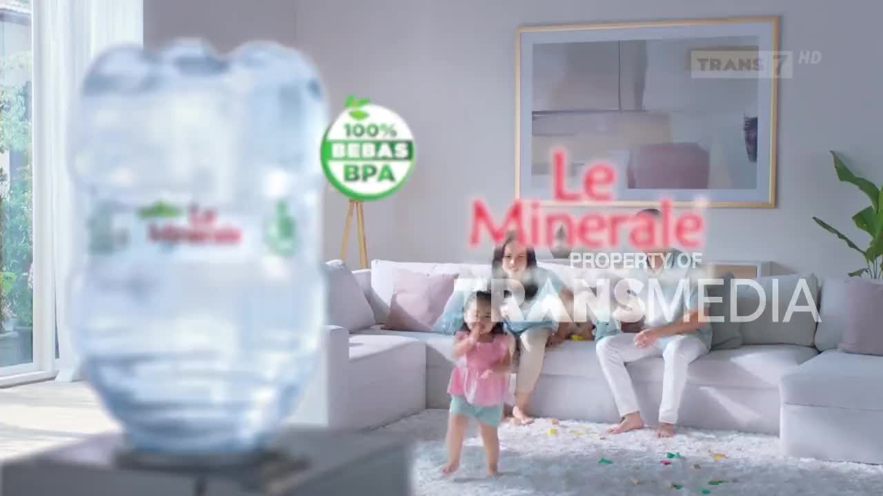 A young girl in a pink top dances on a fluffy rug while her parents watch from the sofa. A large water dispenser stands in the foreground, its label clear.
