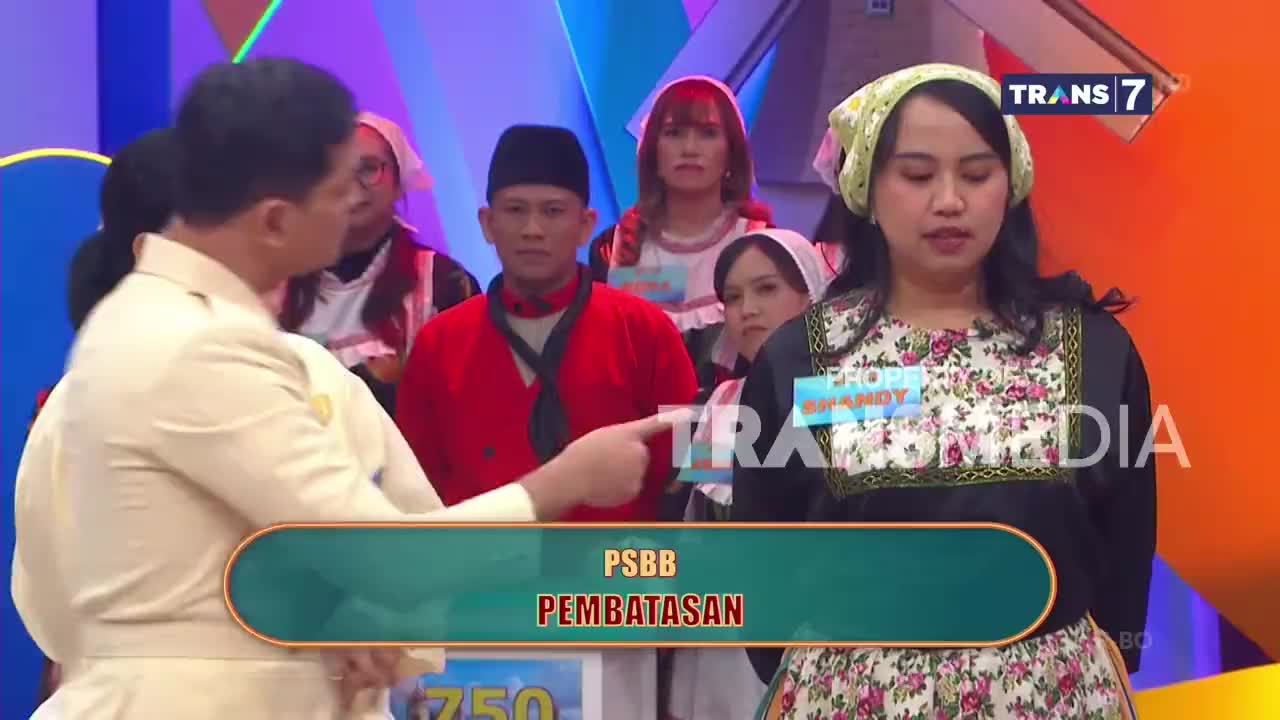 A man in a light suit points his finger towards a woman in a floral dress and headscarf on the Trans7 stage. Behind them, other contestants in traditional Indonesian attire wait their turn.