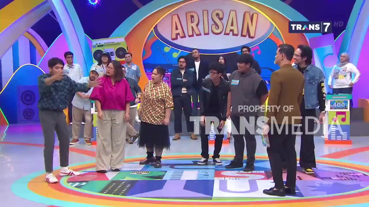 A young man in a patterned shirt gestures emphatically towards a woman in a bright pink top. The two stand on a colorful stage, surrounded by other performers and a banner that reads "ARISAN" in large letters.