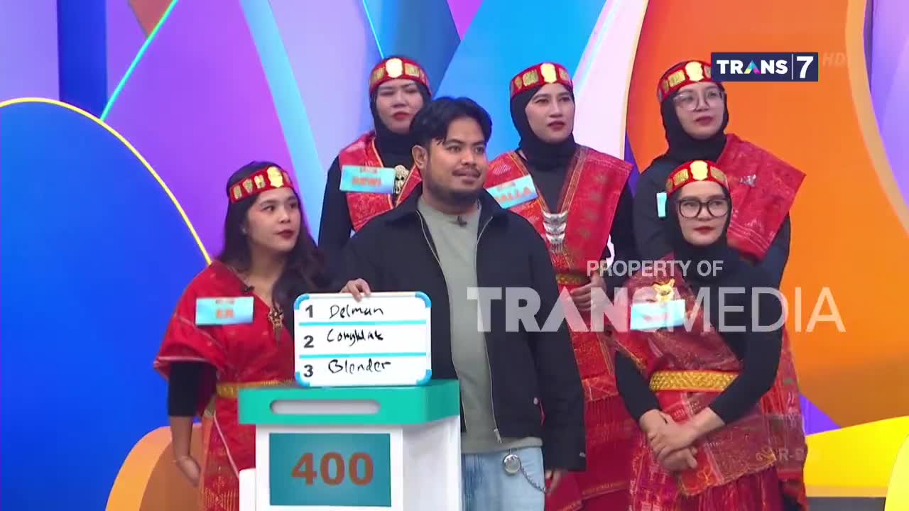 A man holds a whiteboard with a list of words, facing a group of women in traditional Indonesian attire. The Trans7 logo is visible in the background.