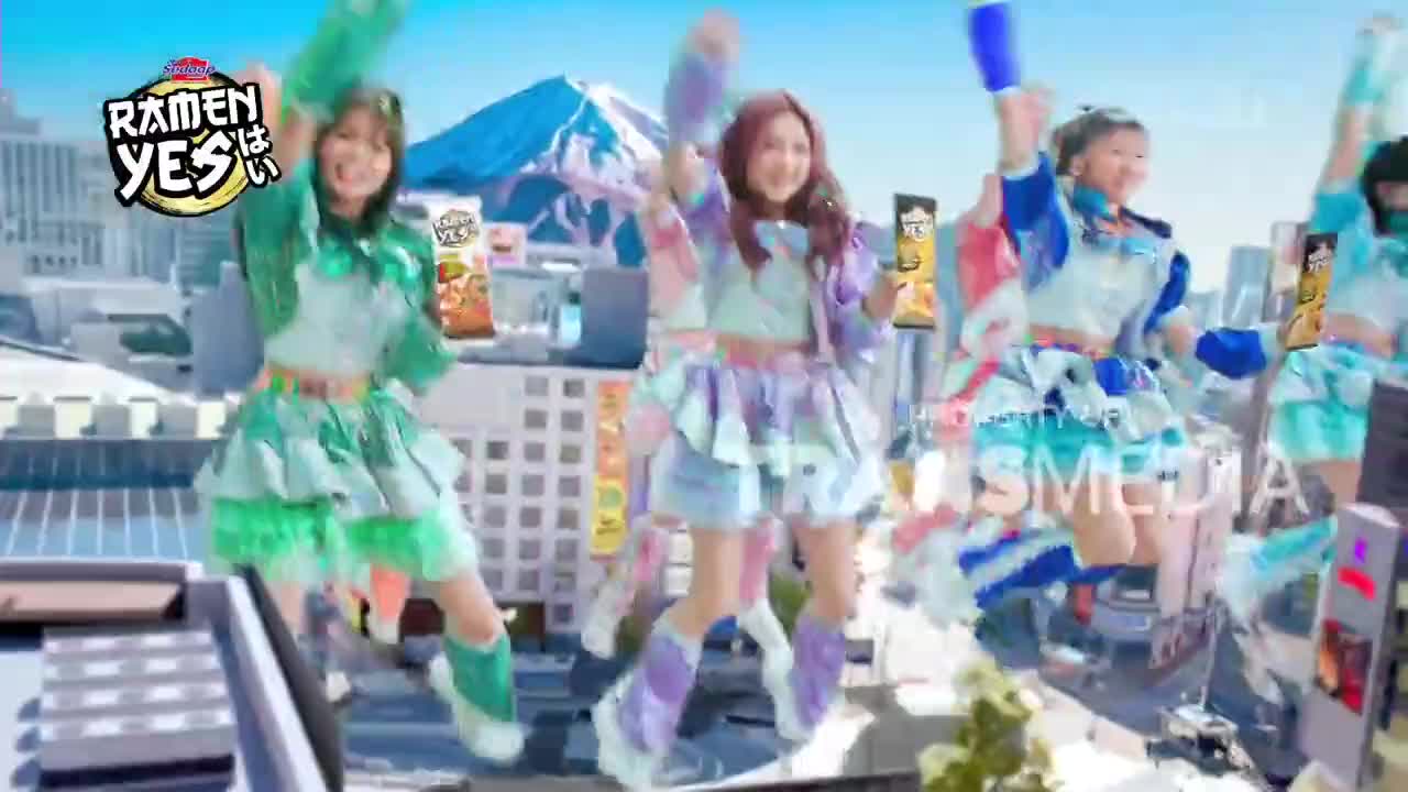 Four young women in brightly colored, layered skirts and jackets are dancing on a rooftop. They are holding up what look like large, colorful candy sticks and smiling. The background features a stylized cityscape with a snow-capped mountain in the distance.