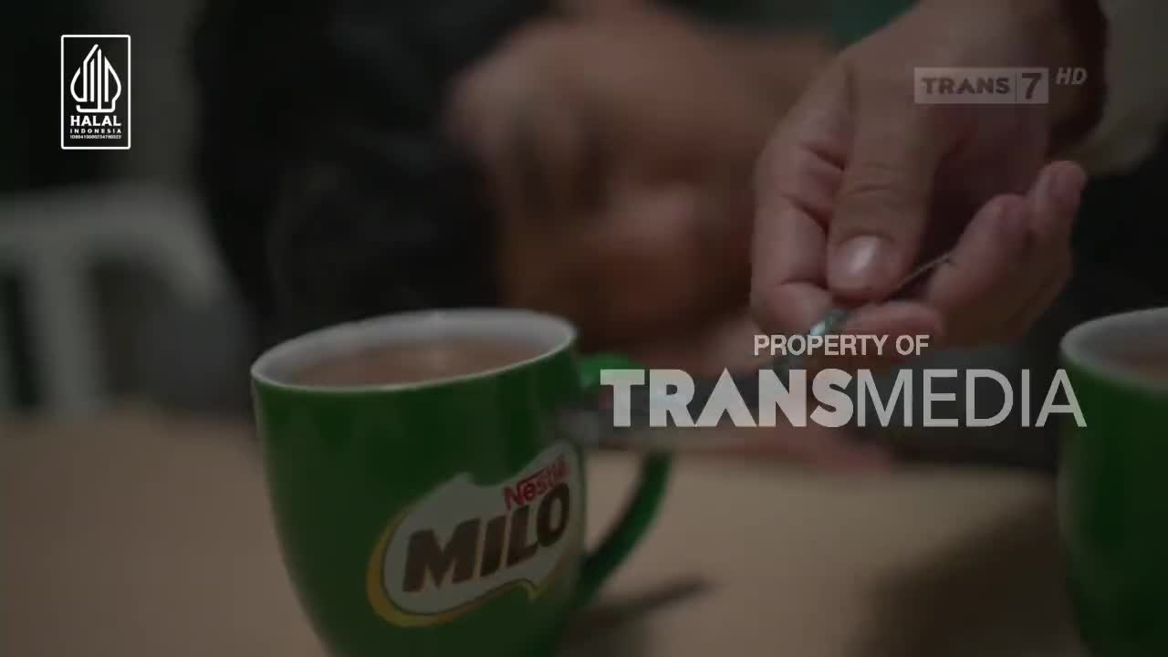A hand carefully stirs a spoon into a mug of Milo, the rich brown liquid swirling. The Trans7 logo is visible in the background, hinting at a broadcast from Indonesia.