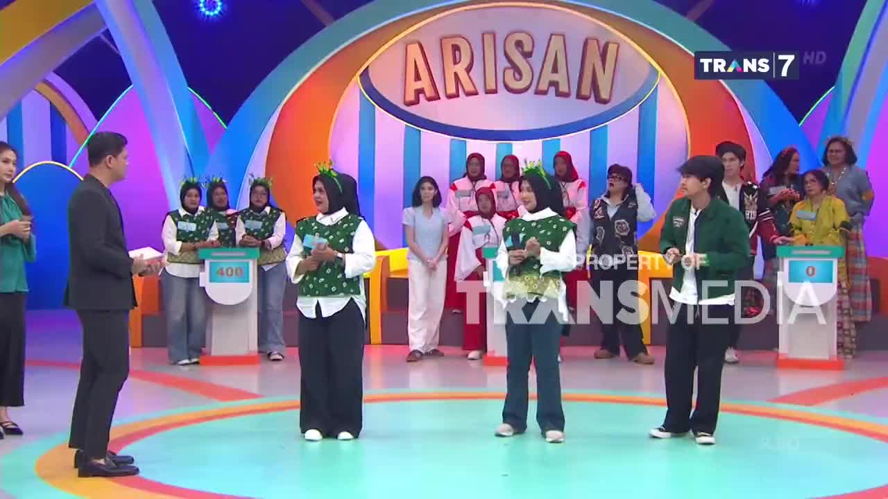 A man in a suit stands facing a group of women on a brightly lit stage, their matching green vests and headbands catching the light. Across the floor, another young person in a green jacket turns towards the center of the action.