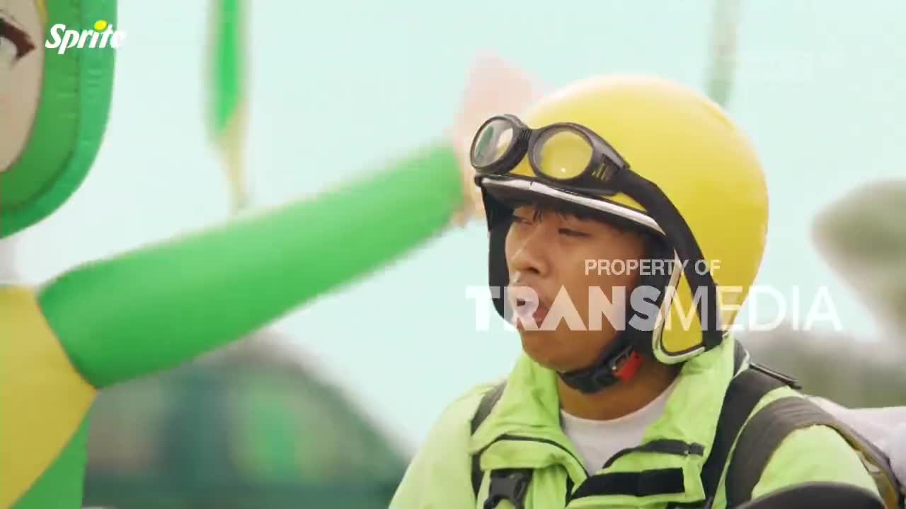 A man in a bright yellow helmet and goggles is focused ahead, his face illuminated by the sun. Beside him, a large green inflatable figure waves its arm.