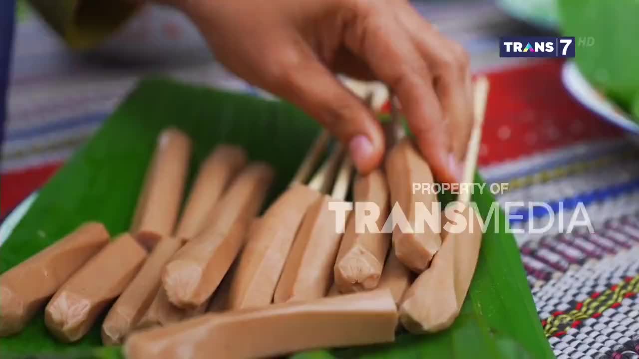 A hand reaches for skewers of sausage laid out on a banana leaf. This preparation is likely for a street food dish, common in Indonesia.