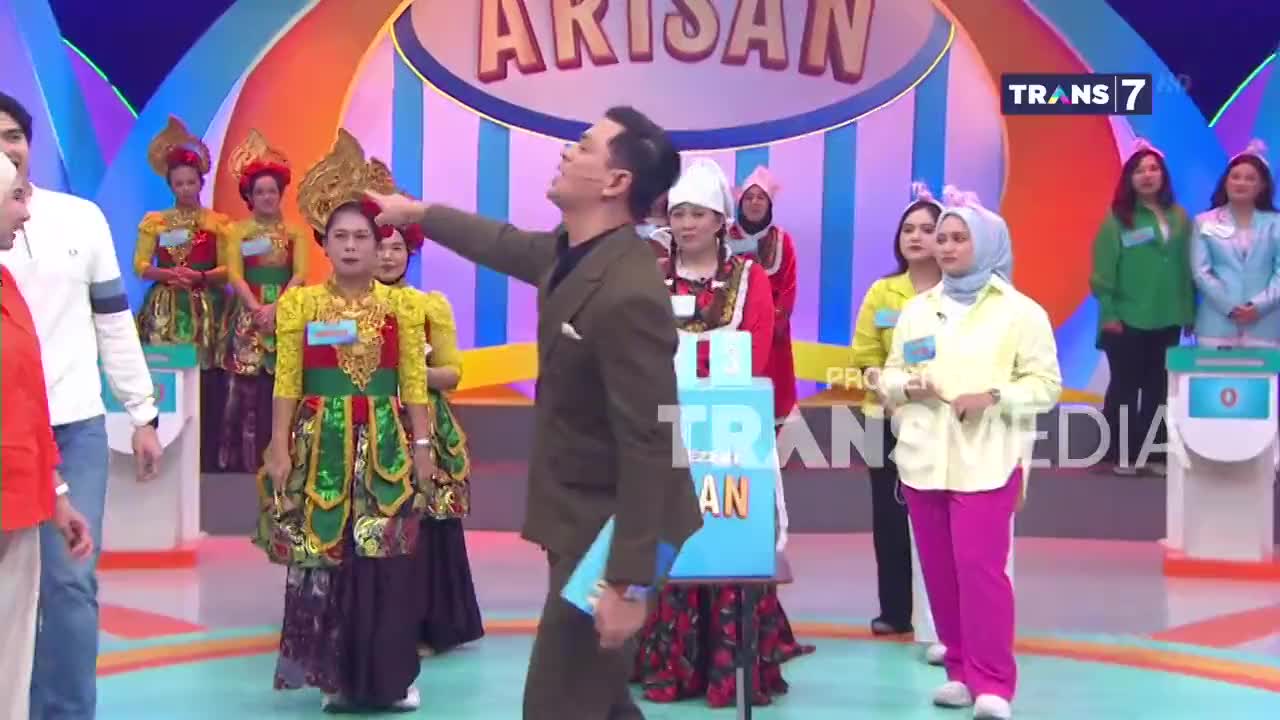 A man in a brown suit points towards a woman in a yellow and green traditional outfit. Several other women in colorful attire stand behind them on a brightly lit stage.