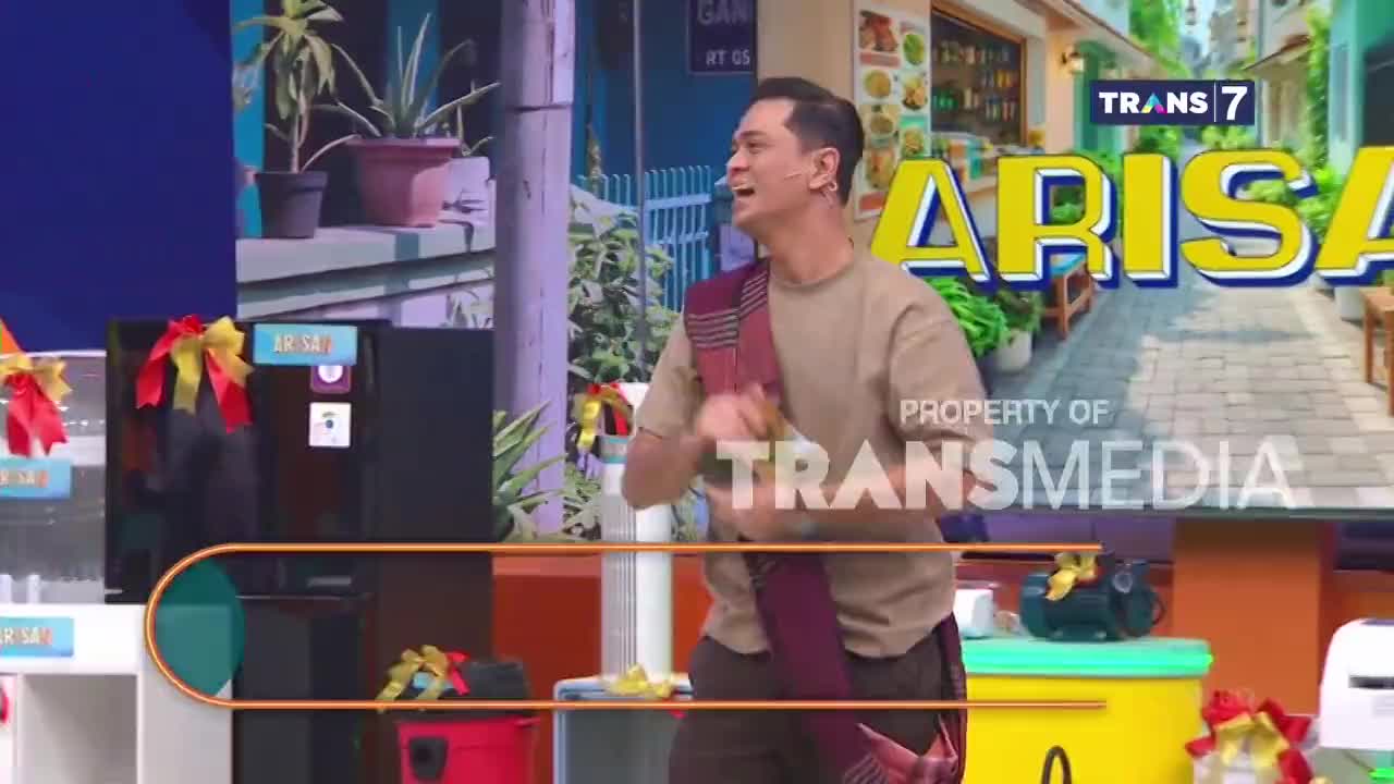 A man in a tan shirt and maroon sash is enthusiastically gesturing on a brightly lit set for Trans7's "Arisan." Behind him, a large black refrigerator is decorated with festive red and yellow ribbons.