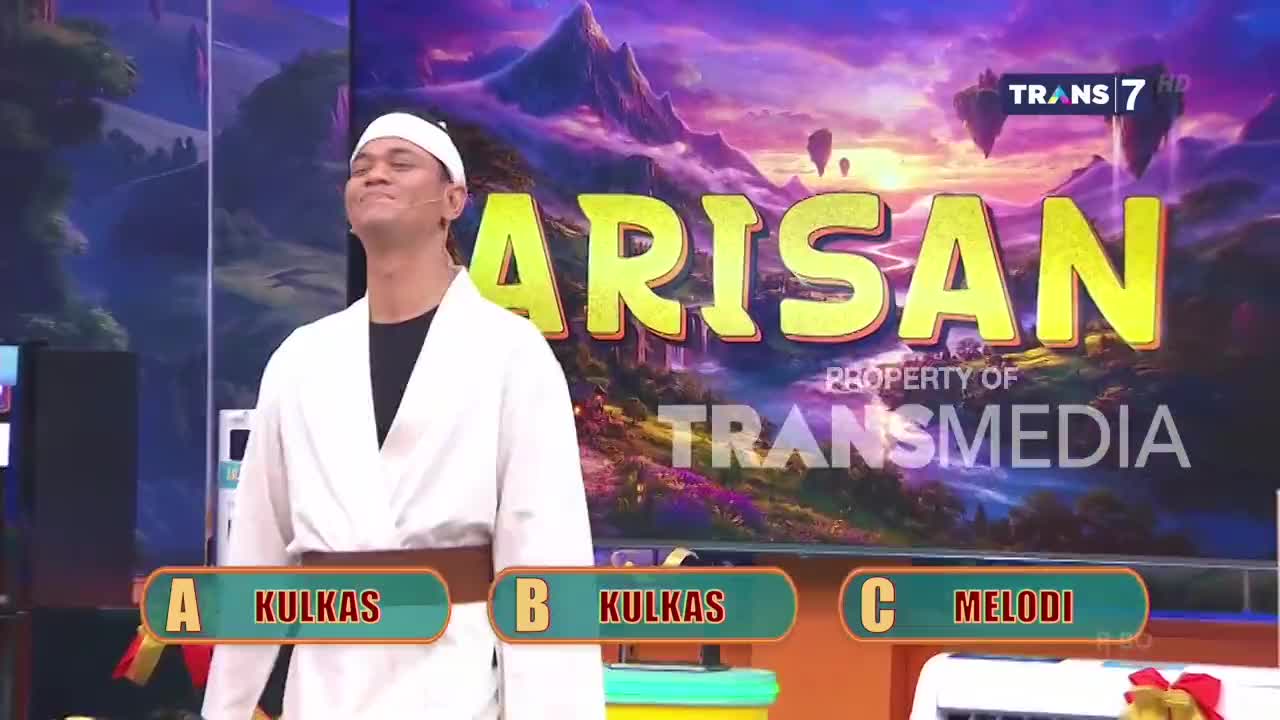 A man in a white gi stands on a brightly lit stage, looking up with a determined expression. Behind him, the Trans7 logo and the word "ARISAN" are displayed against a fantastical landscape.