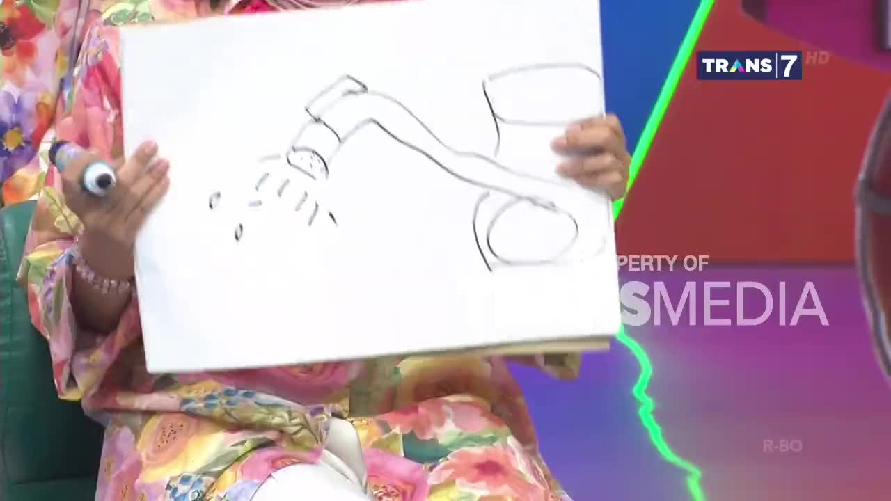 A person in a floral shirt holds up a drawing of a sprinkler, with water droplets falling from it. The Trans7 logo is visible in the background, indicating this is part of a broadcast in Indonesia.