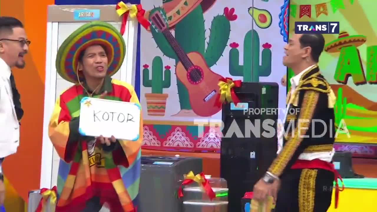 A man in a colorful poncho and sombrero holds up a whiteboard with "KOTOR" written on it. To his right, another man in a matador-like outfit stands, looking towards the audience. The backdrop is a vibrant Mexican-themed set, complete with cacti and a guitar.