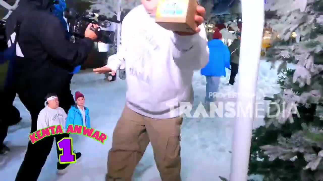 A man in a white hoodie holds up a small box, his arms outstretched. Behind him, a cameraman films the scene as a Trans7 logo appears.