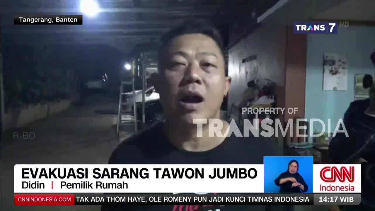 A man speaks directly to the camera, his mouth open as if in mid-sentence. Behind him, a dark alleyway is illuminated by a bright light, and a ladder stands against a wall.