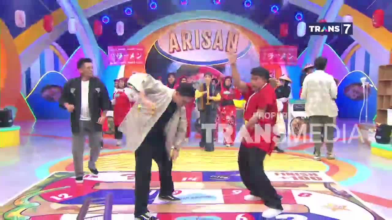 Two men are dancing on a colorful game board, their movements energetic and playful. Behind them, a crowd watches from a brightly lit stage, the "ARISAN" logo prominently displayed above.