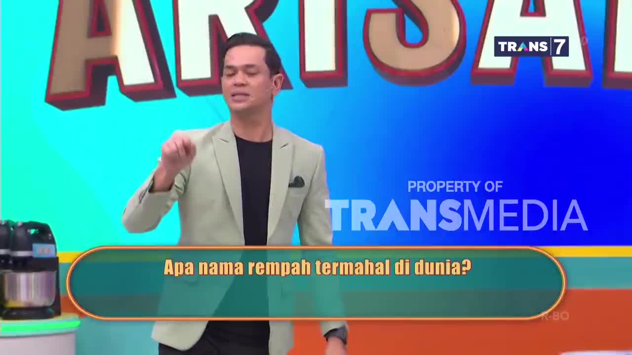 A man in a light suit gestures emphatically as a question appears on a screen: "What is the most expensive spice in the world?" The Trans7 logo is visible in the background.