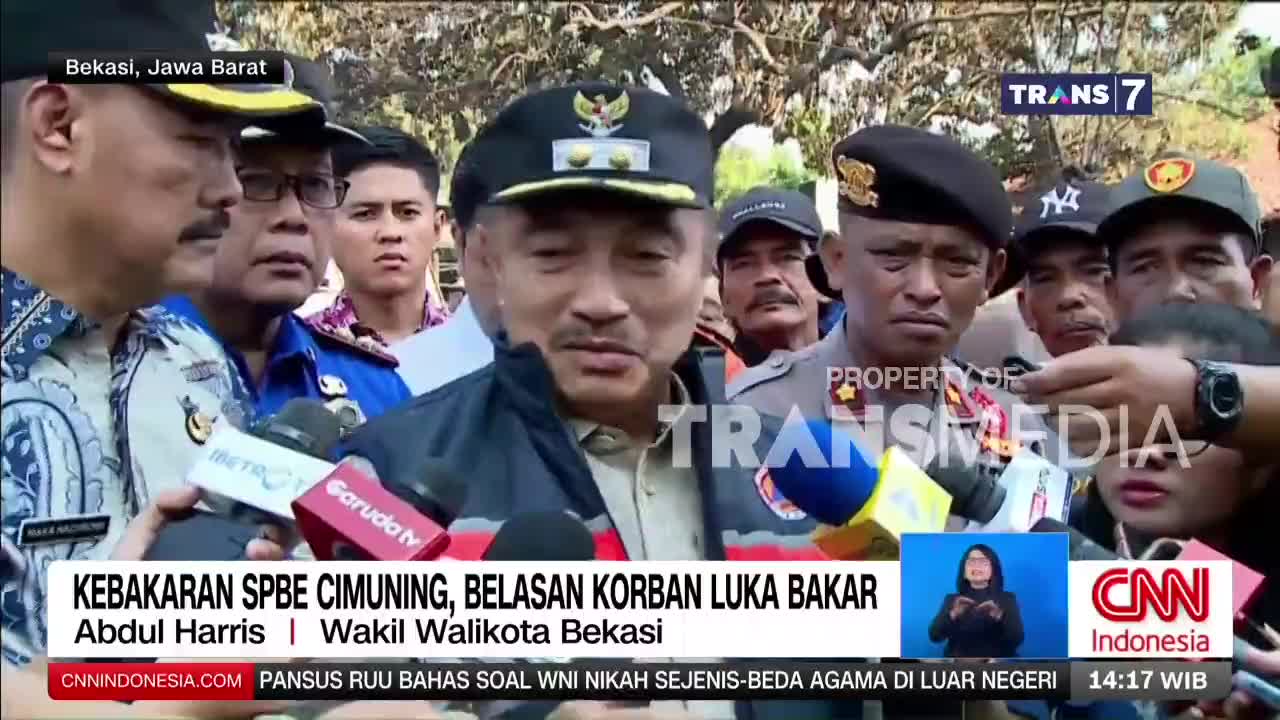 A man in a dark jacket and cap speaks into multiple microphones, surrounded by reporters. The Trans7 logo is visible above the crowd.