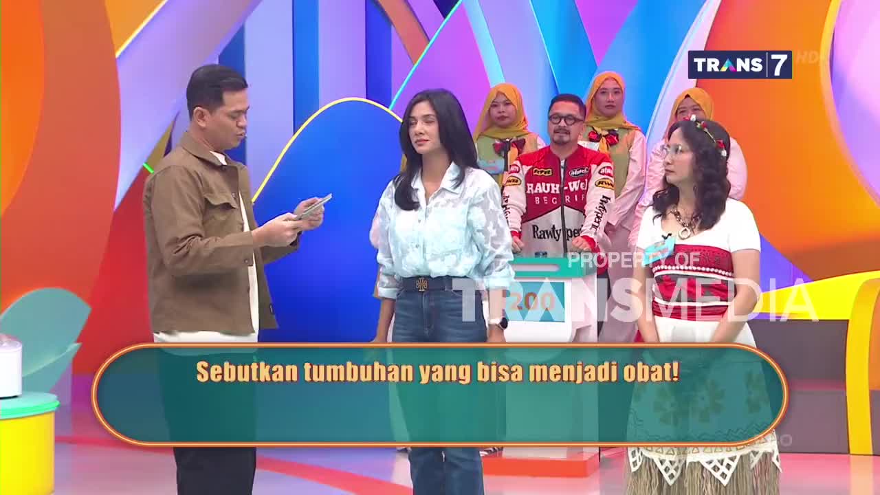 A man in a brown jacket holds out cards to a woman in a patterned shirt. Behind them, a group of people watch from a colorful stage set, with the Trans7 logo visible.