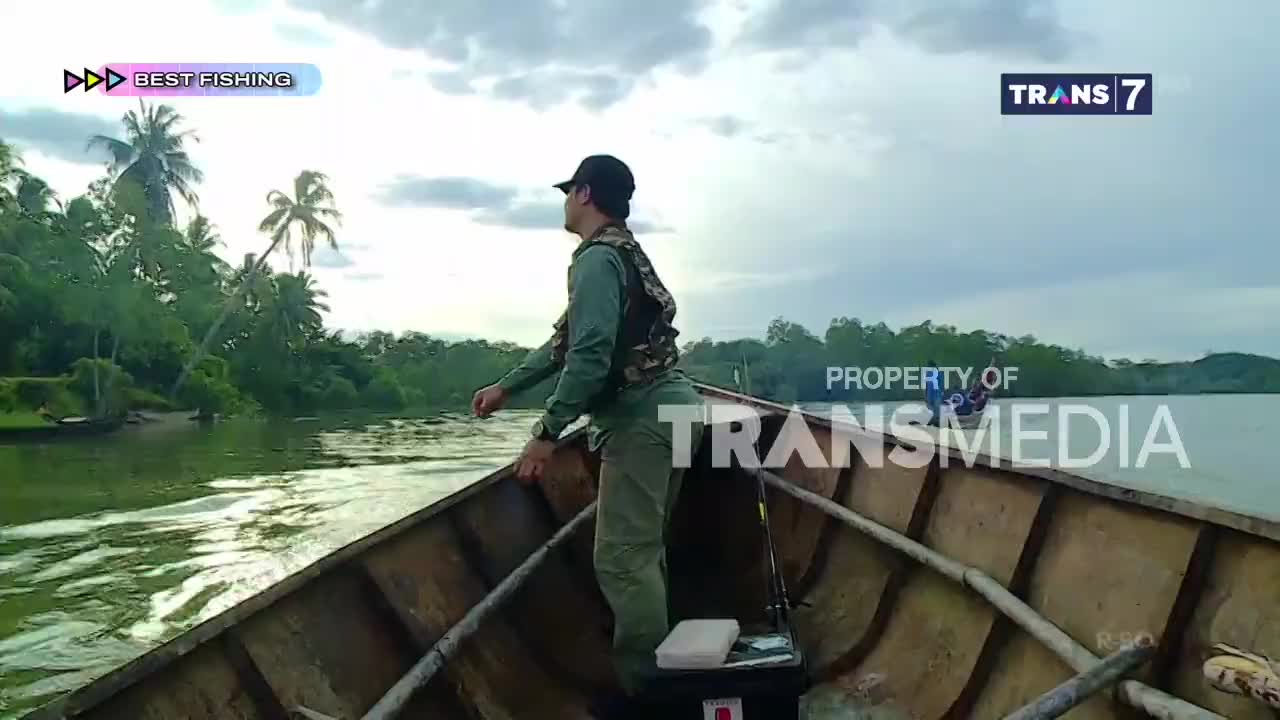 The wooden boat glides through the murky green water, its passenger scanning the horizon. Lush Indonesian foliage lines the riverbanks under a cloudy sky, a scene likely captured for a Trans7 fishing program.
