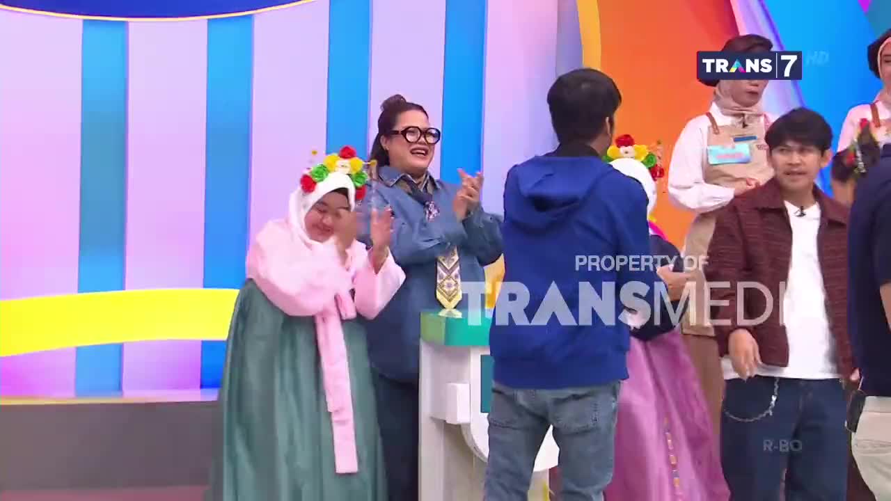 A woman in a pink hanbok and a floral headdress claps her hands, her face turned down. Beside her, a woman in a denim jacket smiles and also claps, her glasses perched on her nose.