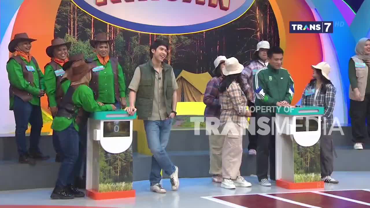A group of contestants, dressed in green shirts and brown vests, stand ready at their stations. A young man in a khaki vest and jeans leans casually against a podium, smiling.