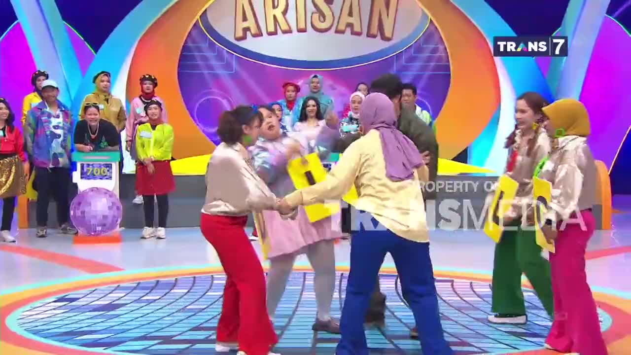 Two women in bright pants are holding yellow signs, pulling against each other. A crowd watches the game unfold on the Trans7 stage.