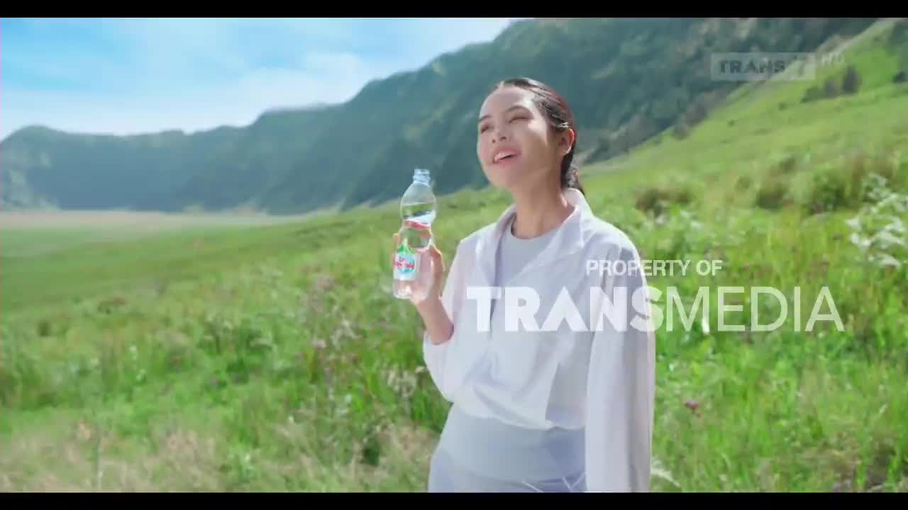 A woman stands in a vast, grassy landscape, holding a bottle of water. The Trans7 logo appears on screen, indicating this is part of an Indonesian broadcast.