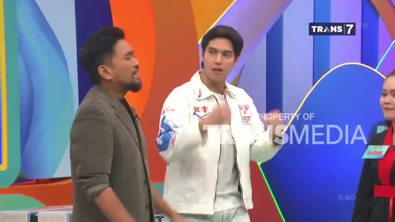 A man in a brown suit turns to speak with a younger man in a white jacket, both on a brightly colored stage. The Trans 7 logo is visible in the background.