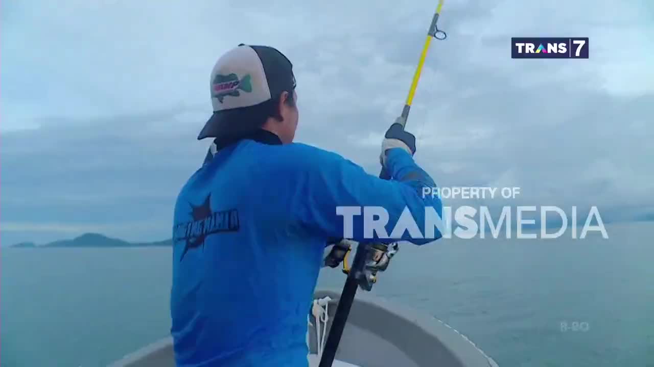 A man in a blue shirt and a fishing hat casts his line from a small boat on the water. The Trans 7 logo appears on screen as he fishes off the coast of Indonesia.