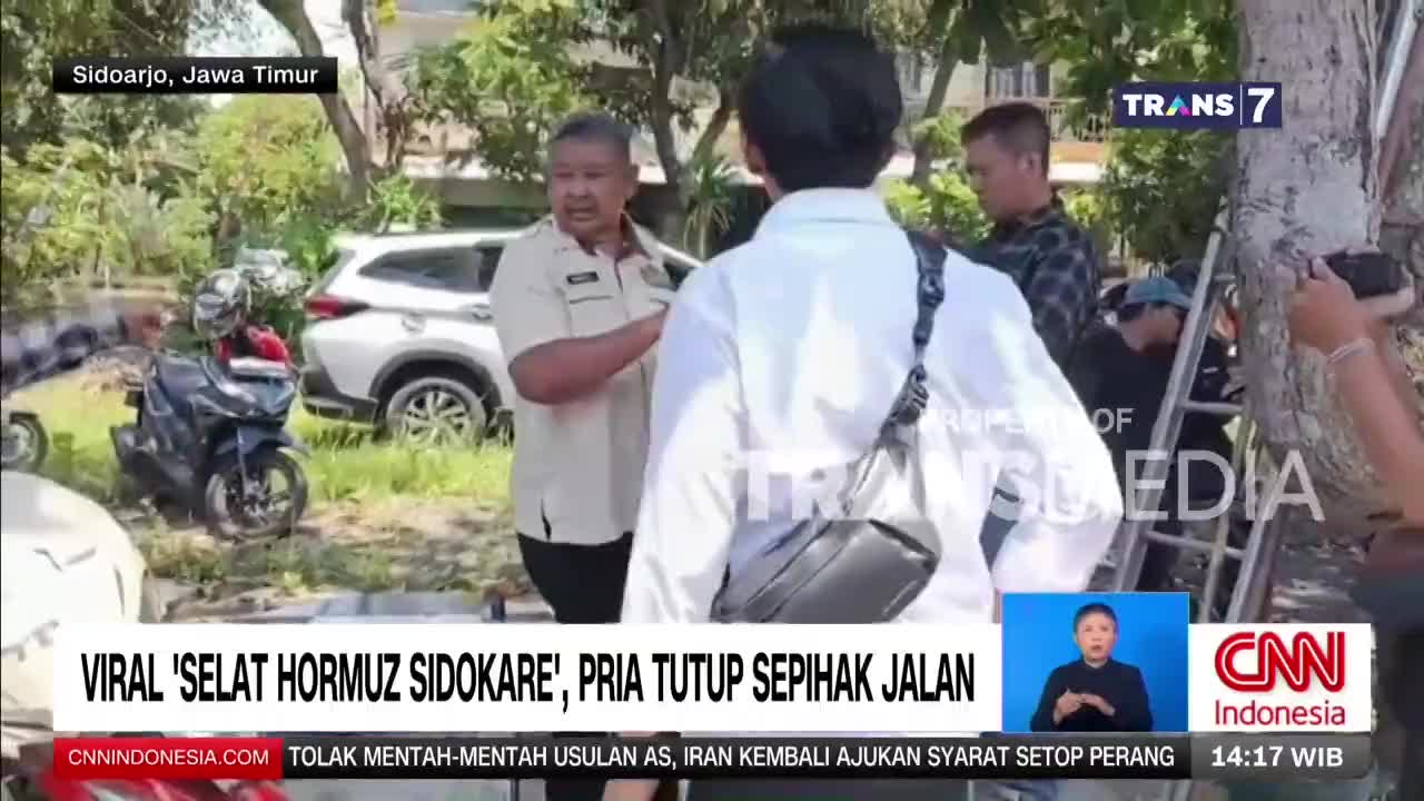 A man in a light brown uniform speaks with someone in a white shirt, while a motorcycle sits nearby. A Trans7 logo is visible in the upper right corner.