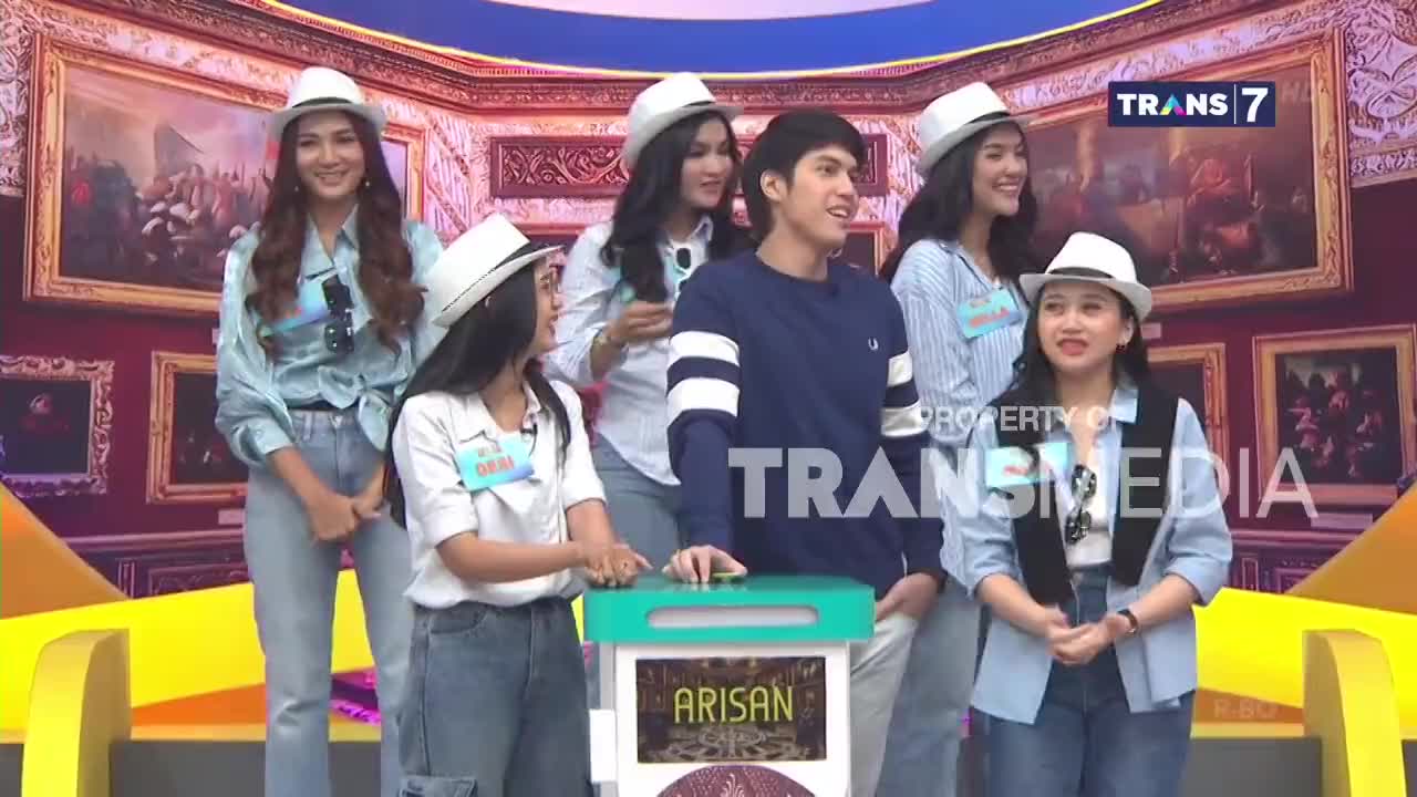 A group of young people, some wearing white hats, stand on a colorful stage in front of framed paintings. The Trans7 logo is visible, and a box labeled "ARISAN" sits on a stand. A group of young people, some wearing white hats, stand on a colorful stage in front of framed paintings. The Trans7 logo is visible, and a box labeled "ARISAN" sits on a stand.