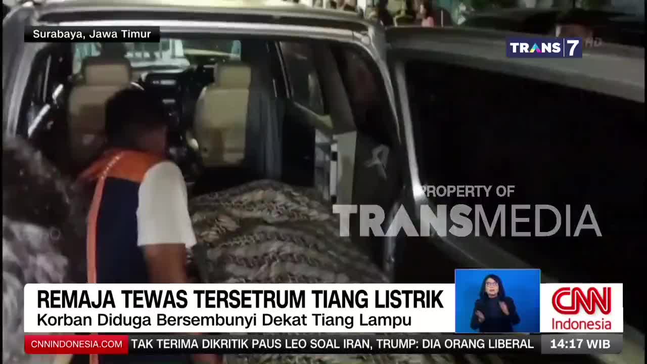 A man in an orange vest carefully places a patterned blanket into the back of an open vehicle. The Trans7 logo is visible on the side of the car.