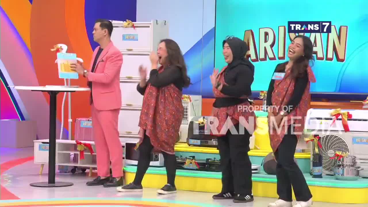 A man in a bright pink suit holds a duck-shaped prop while three women in patterned wraps clap and laugh on the Trans7 stage. The women's joyful expressions suggest they're enjoying the game or segment.