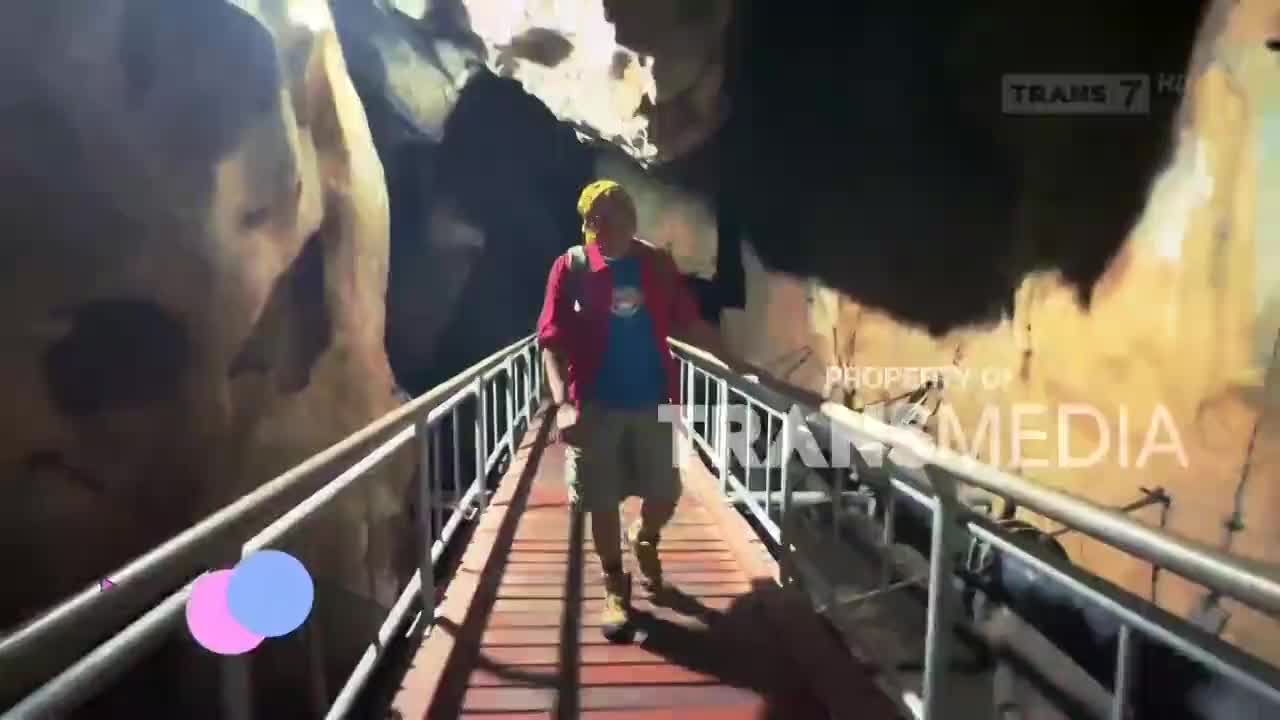 A man in a red jacket and yellow bandana walks towards me on a narrow walkway inside a cave. The Trans7 logo is visible on the screen.