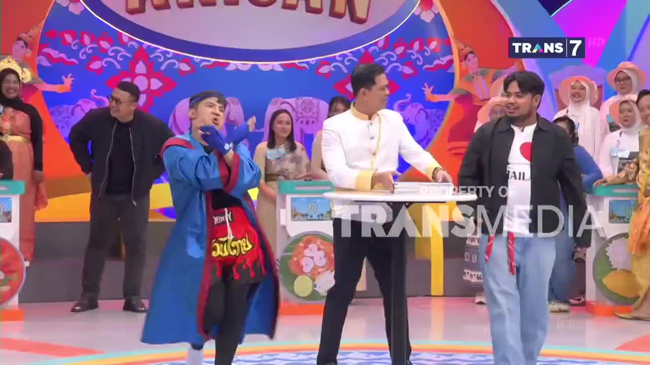 A performer in a blue robe and red shorts strikes a pose on the Trans7 stage. Beside him, a man in a white jacket holds a tray, while another man in a black jacket watches.