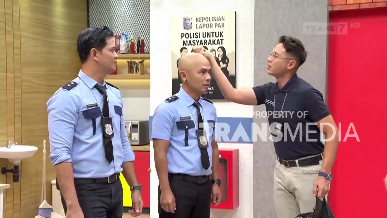 A man in a dark polo shirt playfully pats the head of another man in a light blue uniform. The third man in uniform watches them, standing to the left.