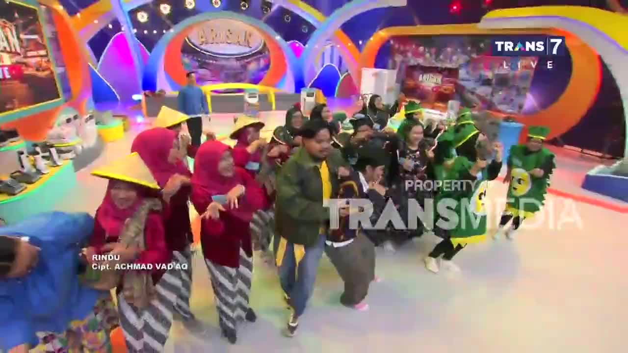 A group of dancers in conical hats and red tops sway rhythmically on the colorful studio floor. Off to the side, two performers in green costumes with yellow smiley faces bounce in unison.