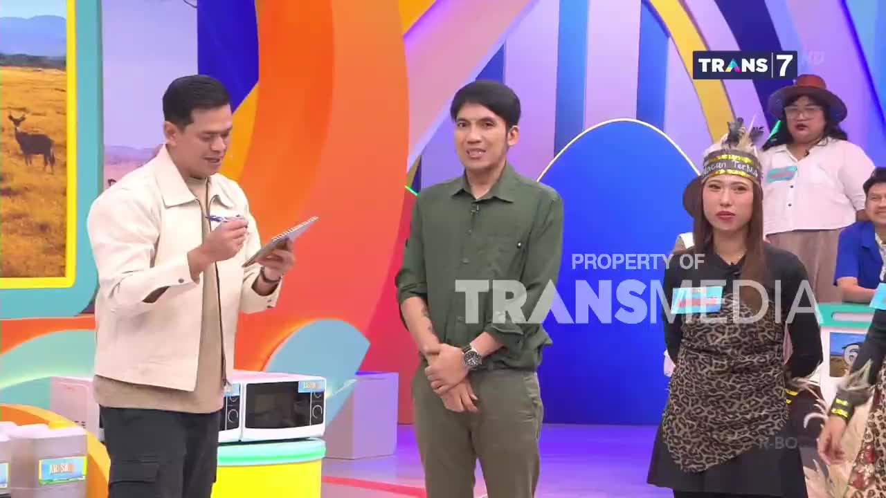 A man in a cream jacket holds a tablet, pen poised. A man in a green shirt stands facing him, hands clasped. Behind them, a woman wears a feathered headband and a leopard-print apron, participating in a Trans7 broadcast.