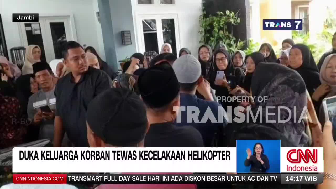 A crowd of people, many dressed in black, are gathered outside a building. The Trans 7 logo is visible in the upper right corner, and a banner at the bottom reads "DUKA KELUARGA KORBAN TEWAS KECELAKAAN HELIKOPTER," which translates to "Grief of the family of the helicopter crash victims."